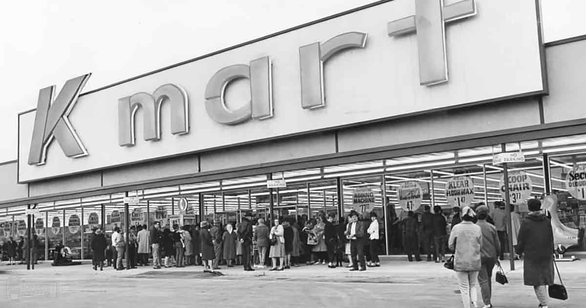 An adult male singer in a mid-20th-century Kmart store, winter setting, clear view into interior with products displayed on shelves, muted tones typical of black-and-white photography from the era.