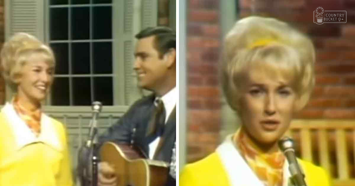 A woman in a yellow and white outfit sings into a microphone on stage with an adult male guitarist playing guitar, both dressed for warm weather, under artificial lighting.