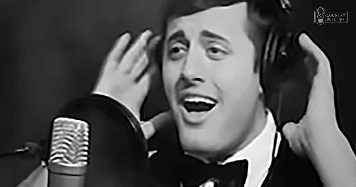 An adult male performer in a black tuxedo with white shirt and black bow tie, likely at a recording studio or stage setting.