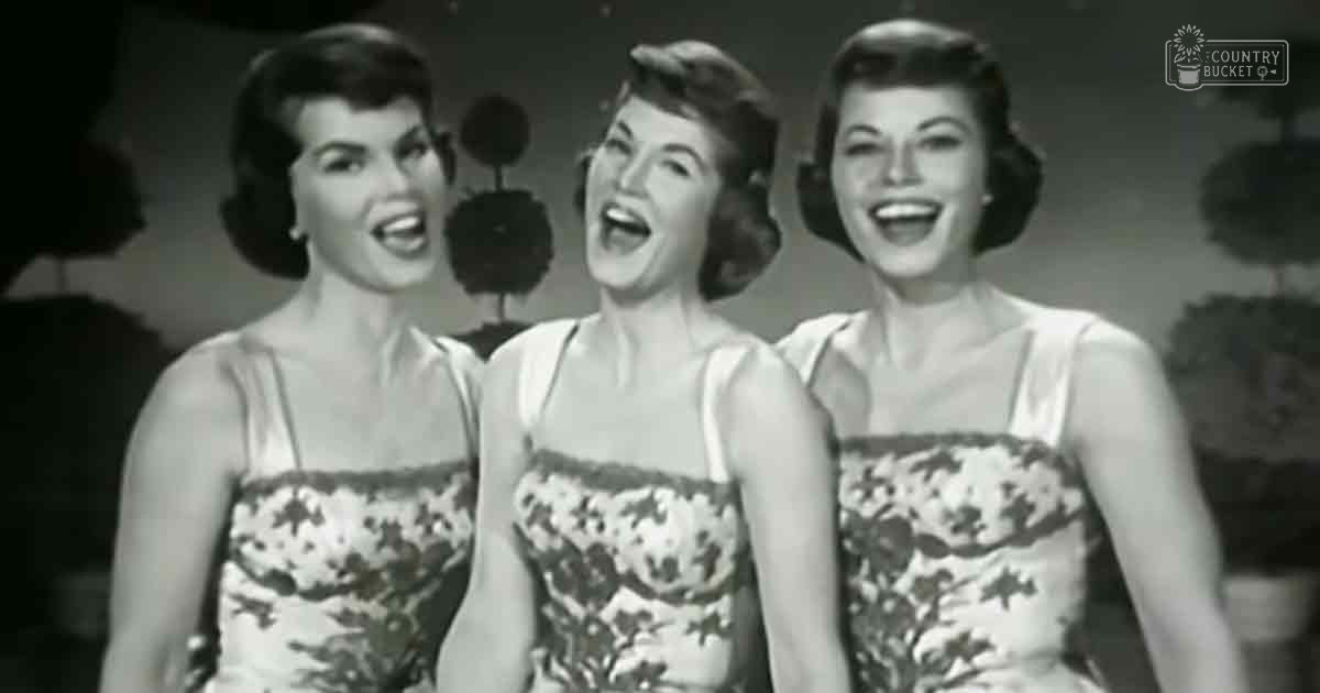 A group performance scene from an older era, mid-20th century, with young adult female performers in form-fitting strapless dresses with floral patterns and white bases. Setting appears to be indoors on a stage, likely during a well-lit indoor performance.