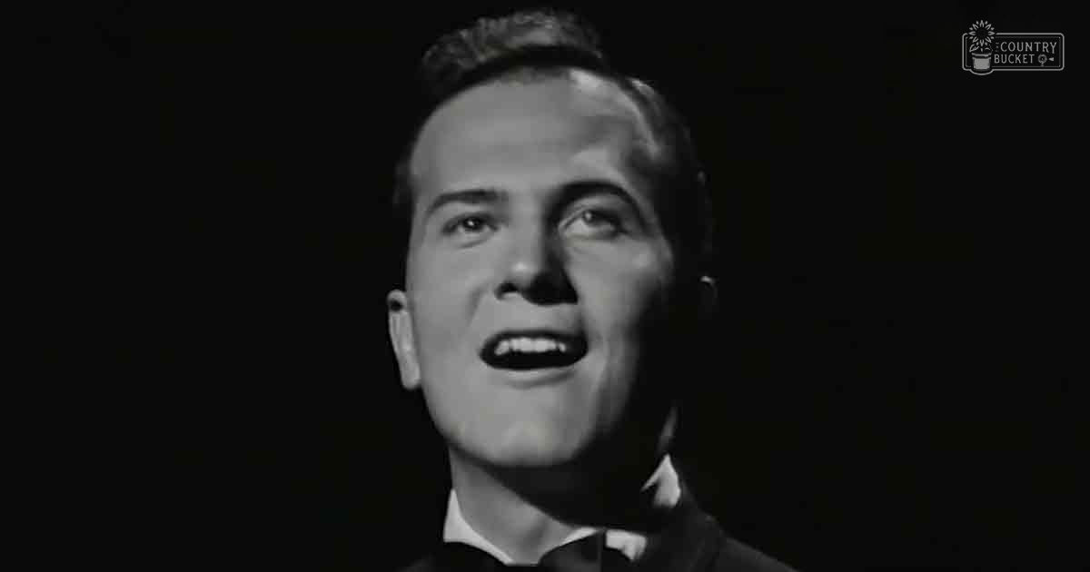 A young adult male in a formal black suit jacket, white shirt, and dark tie from mid-20th century indoors with dramatic lighting at night.