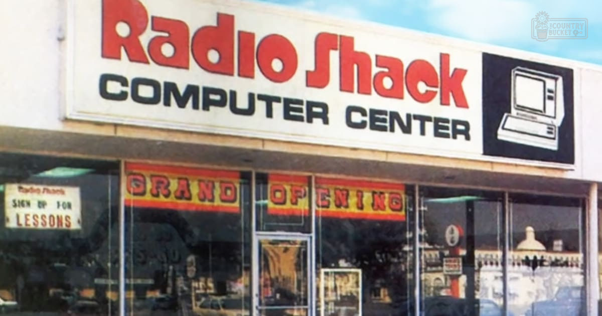 Urban storefront of a Radio Shack Computer Center with large windows displaying 'GRAND OPENING' signs, with prominent red signage against white and black text. Interior suggests furniture and possibly electronic displays.