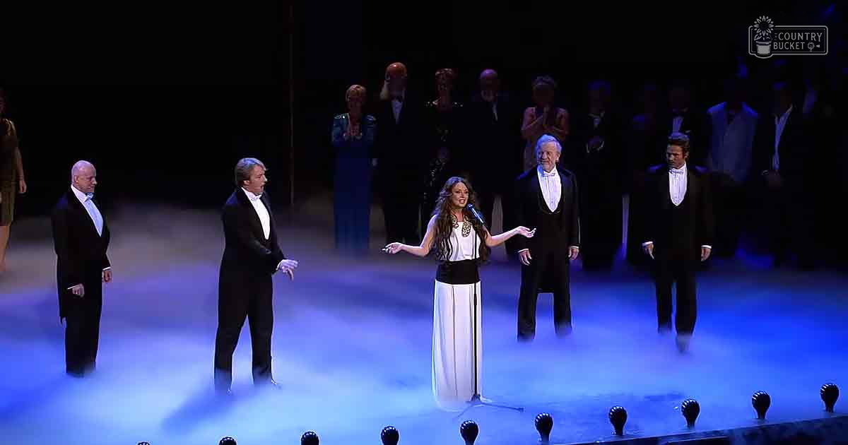 An adult female singer in a white and black dress with high neckline and long sleeves, accompanied by middle-aged and older male performers in formal attire on stage under blue lighting at an opera or classical music performance venue.