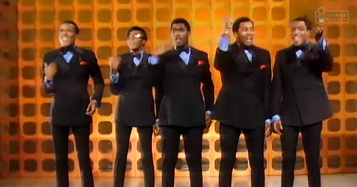 Group performance scene with five adult males in matching black suits, light blue shirts, and red pocket squares on an indoor stage or studio set against a patterned orange backdrop. Bright artificial lighting illuminates the scene.