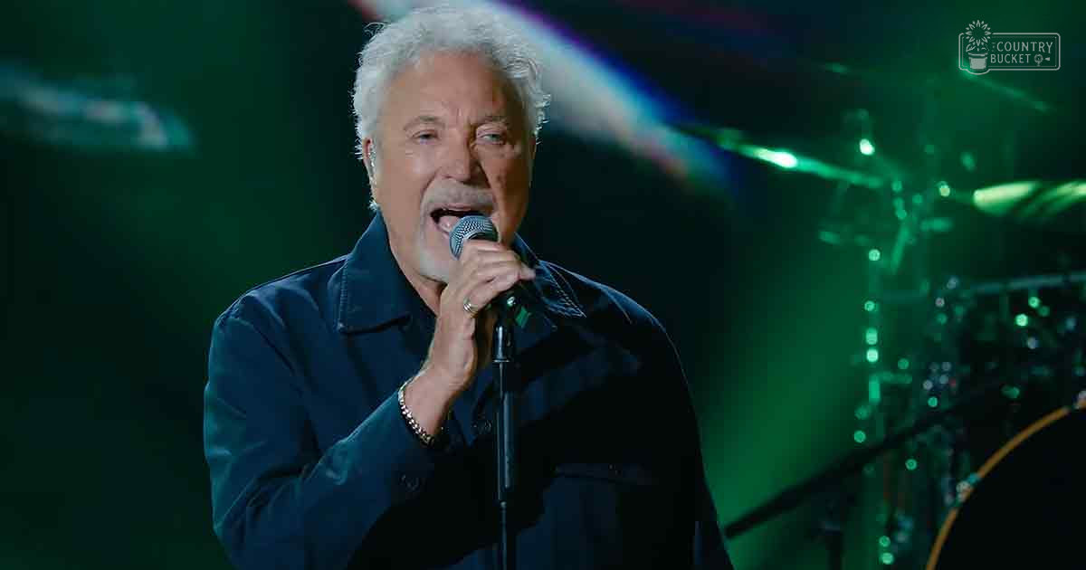 A middle-aged man in a relaxed fit, long-sleeved dark blue shirt performs on stage with modern casual attire under indoor concert venue lighting.