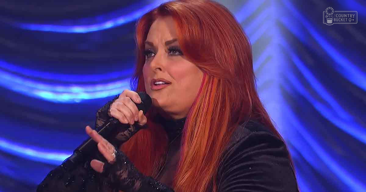 Adult female performer wearing black lace gloves singing into a microphone in a studio setting with blue abstract-patterned background lighting.