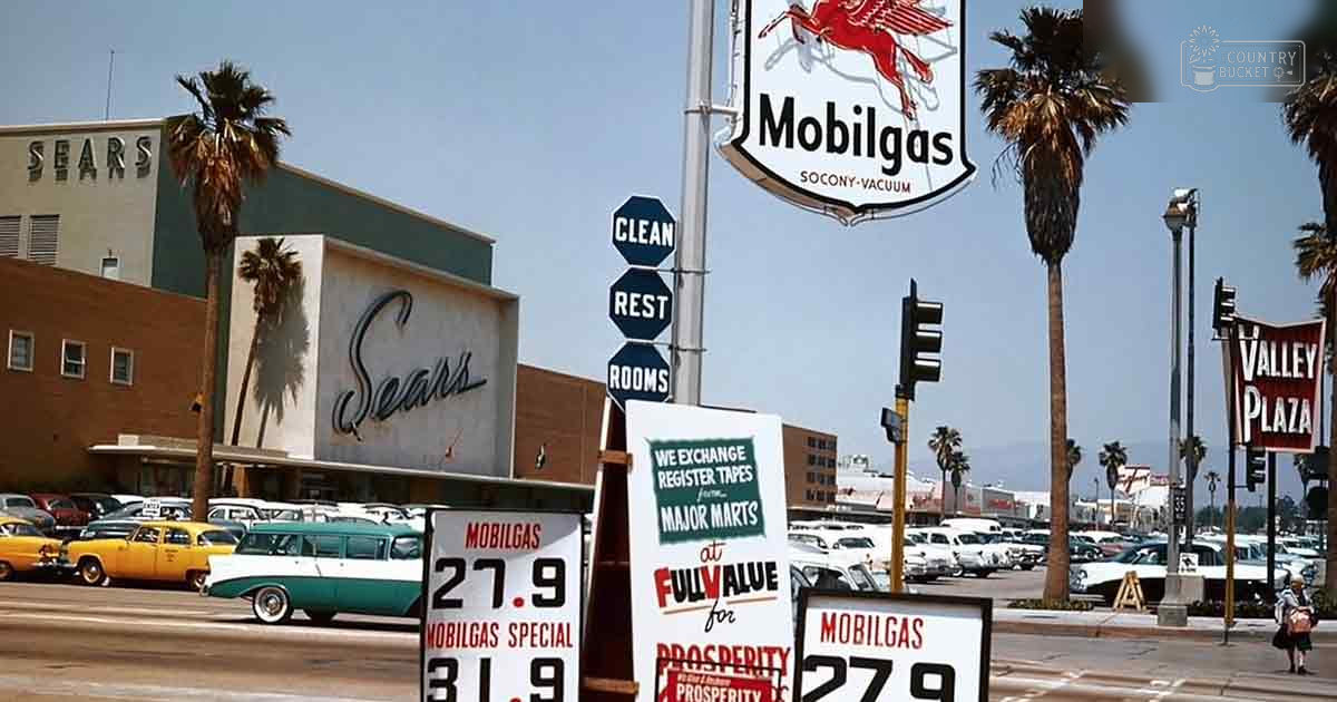 Mid-20th-century American shopping plaza with Mobilgas station signage, palm trees, vintage cars, and a Sears building in a warm climate setting.