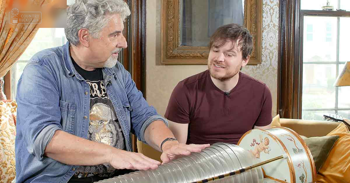 Middle-aged man in a denim jacket with a skull graphic shirt, interacting with an ornate wooden drum in a cozy living room setting.