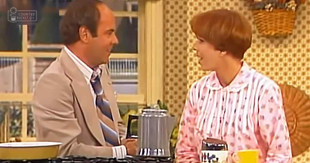 Two individuals in a kitchen setting, one wearing a light beige suit jacket with blue and brown striped tie, another in a pink blouse with white vertical stripes. Polka-dotted wallpaper and woven basket on the wall add to the homey decor.