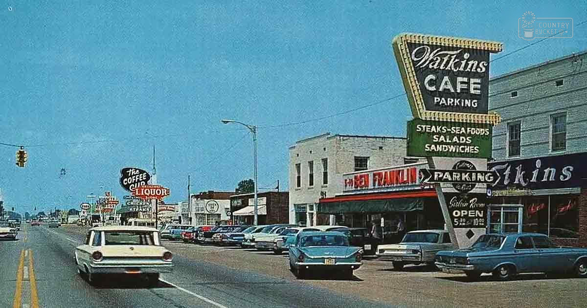 Classic cars from the 1950s-60s parked along a street in Watkins Cafe Parking area, with brightly colored signs for cafes and liquor stores. A clear blue sky suggests daytime during summer or early fall.