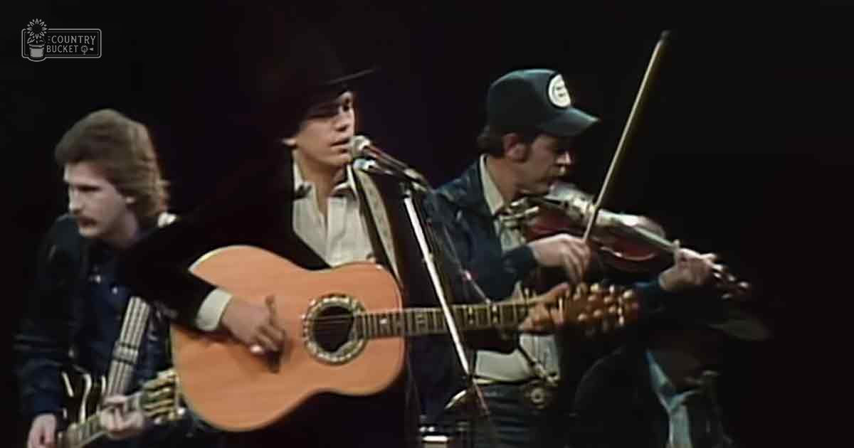 Adult male performers in a 1970s or early 1980s musical performance on stage, wearing jackets and caps under dramatic lighting.