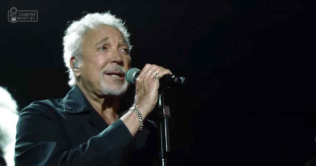 An older adult male singer performing on stage in a dark-colored suit jacket with a V-neckline, surrounded by typical performance equipment and under spotlight lighting.