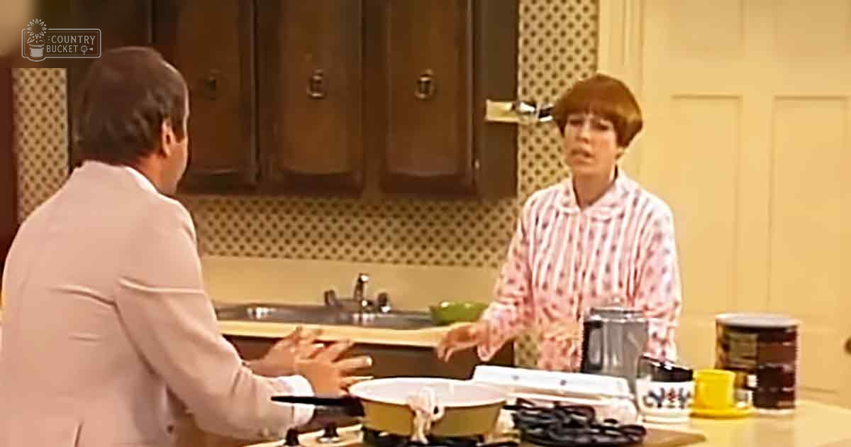 An adult male in a light-colored suit jacket gestures while explaining something near an adult female wearing pink and white striped pajamas, both situated within a kitchen setting with wooden cabinets and patterned backsplash.