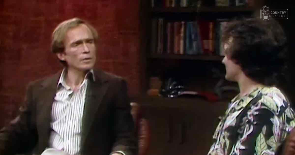 Middle-aged male and young adult female in casual attire against a red textured wall with bookshelf and chair.