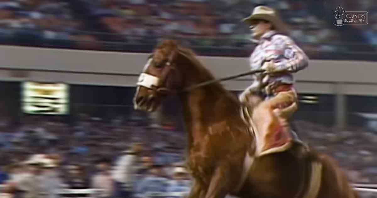A quarter horse breed, likely an adult male rider wearing a wide-brimmed straw hat and plaid shirt in a brightly lit indoor arena during what appears to be a rodeo event.