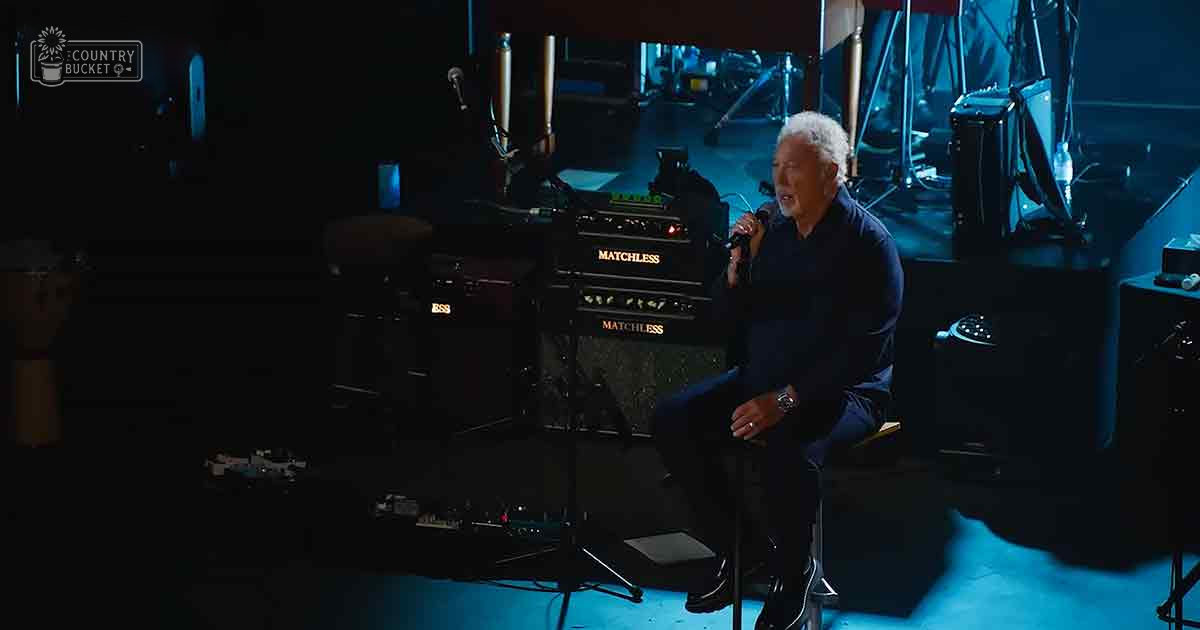 A middle-aged adult male singing into a microphone in a music studio or recording session, surrounded by professional recording equipment under artificial blue lighting.