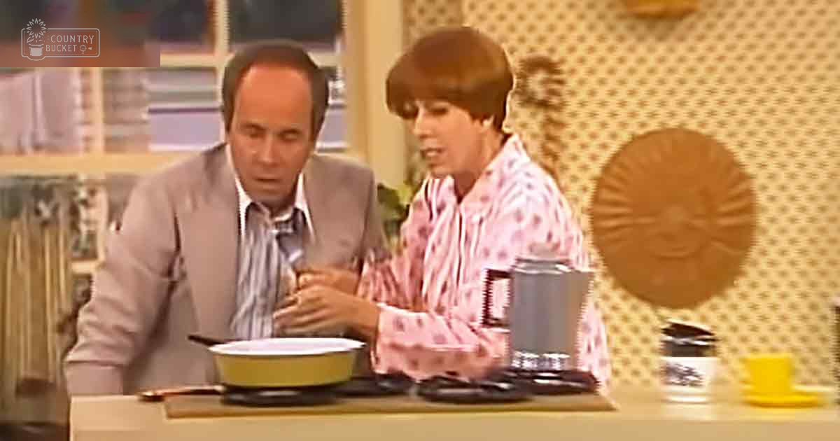 A mid-20th-century kitchen scene with two adults, one wearing a light gray suit jacket over a striped shirt and tie, and the other in a pink and white patterned blouse. The setting includes a small portable stove, coffee maker or thermos, yellow cup and saucer, and simple decor typical of that era.
