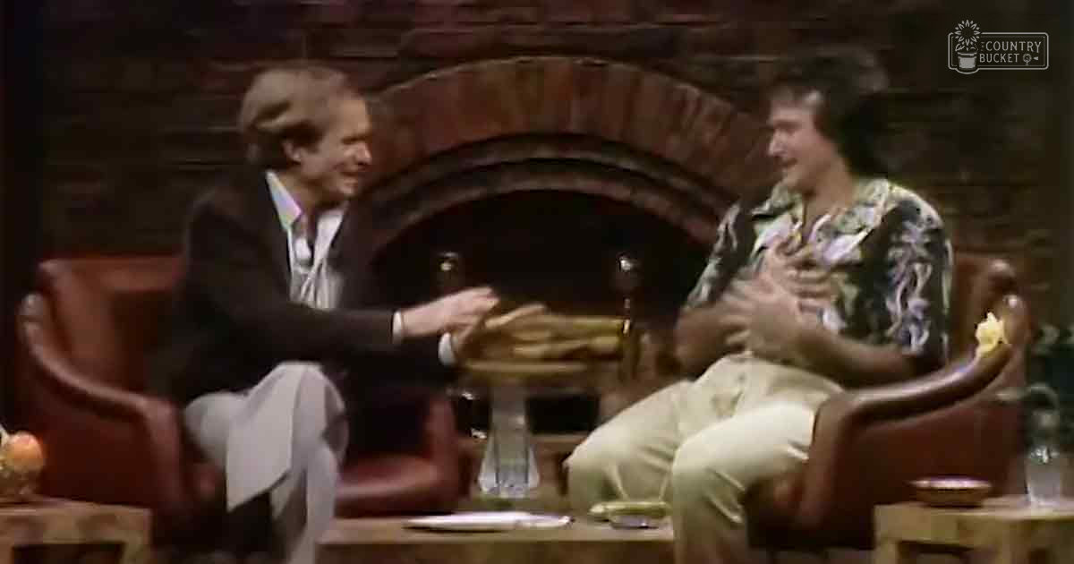 Two adults in a cozy indoor talk show setting with red leather armchairs, a wooden coffee table, and a dimly lit evening ambiance.