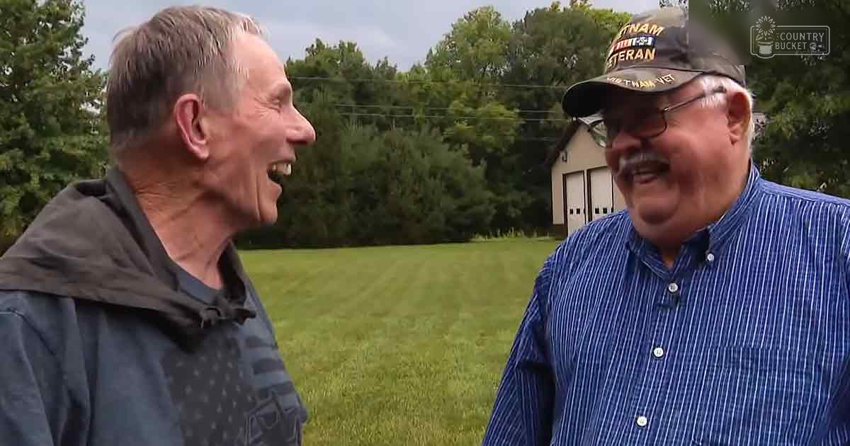 Two older adults wearing casual outdoor clothing in a rural grassy field with trees and part of a building in the background; spring or summer season, late afternoon soft natural lighting