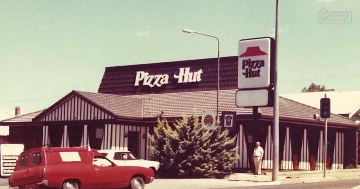 A red van with a white stripe parked in front of a Pizza Hut restaurant in an urban area during daytime under clear skies, likely spring or summer.