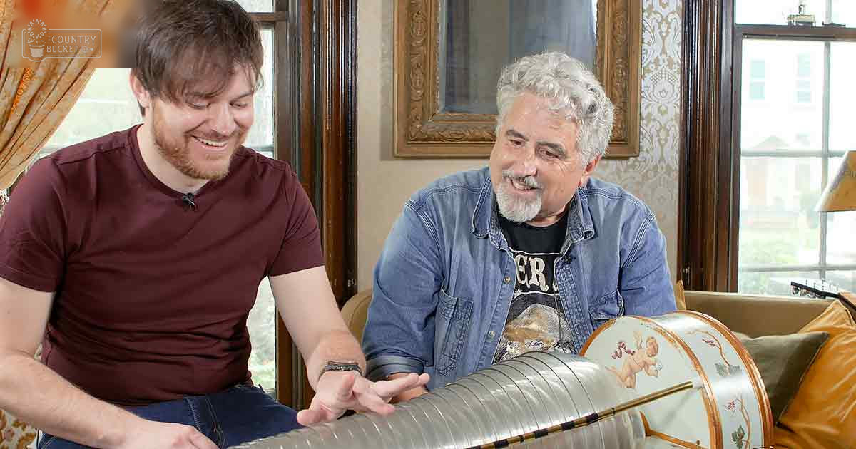 Two adults, one wearing a maroon t-shirt with relaxed fit and crew neck; another older adult in a blue denim jacket over a black graphic tee, interacting near an orange-cushioned couch. A decorative drum with gold accents is prominently featured.