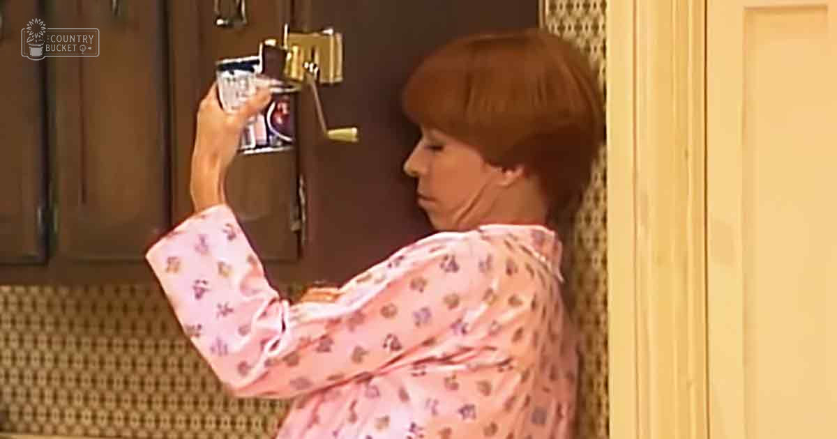 A middle-aged woman in a pink floral-patterned robe stands indoors, holding a glass of water near a door handle with patterned wallpaper and wooden cabinetry visible behind her.