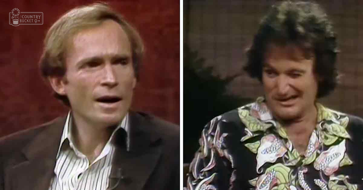 Two adults, a male wearing a dark blazer over a white and black striped shirt with a collar, and another adult male in a loose-fitting floral patterned shirt on a red backdrop in what appears to be a television studio or talk show environment.
