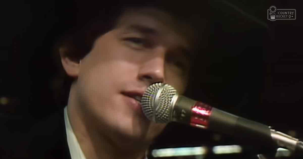 A young adult male singing into a microphone at an indoor event, wearing a white collared shirt with a dark jacket over it.