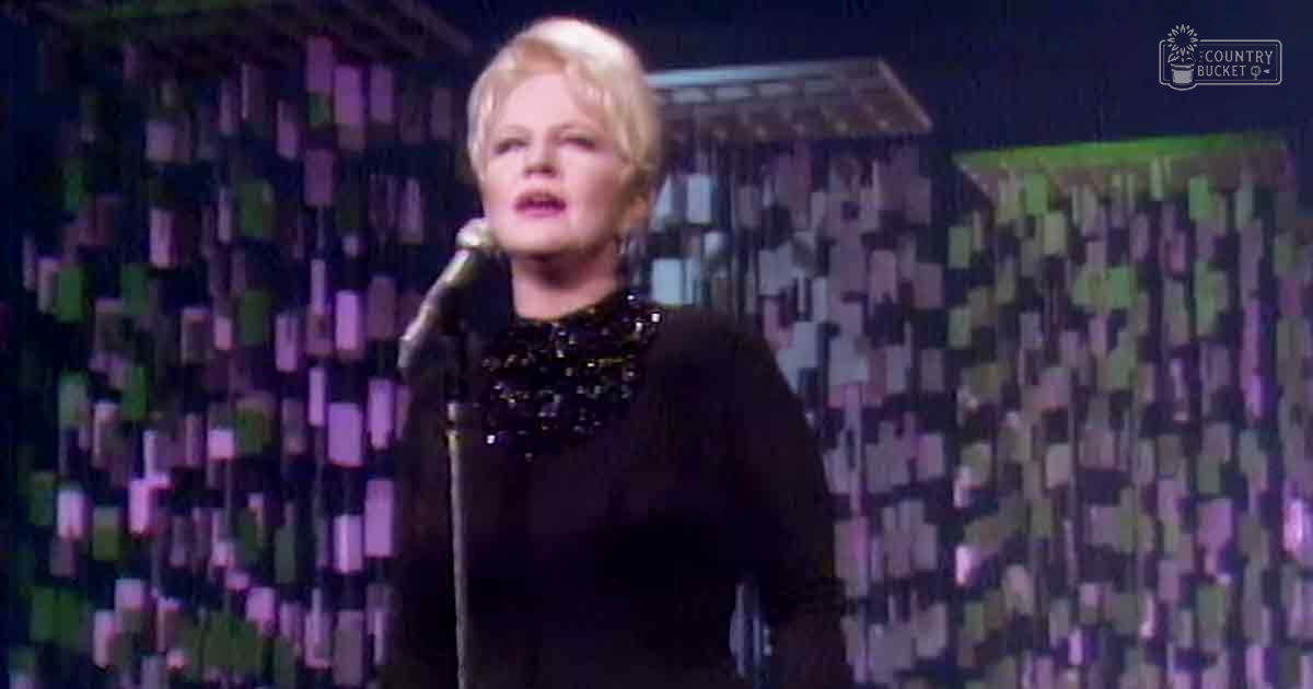 A female adult singer wearing a black sequined dress, holding a microphone on stage with patterned wall backdrop.