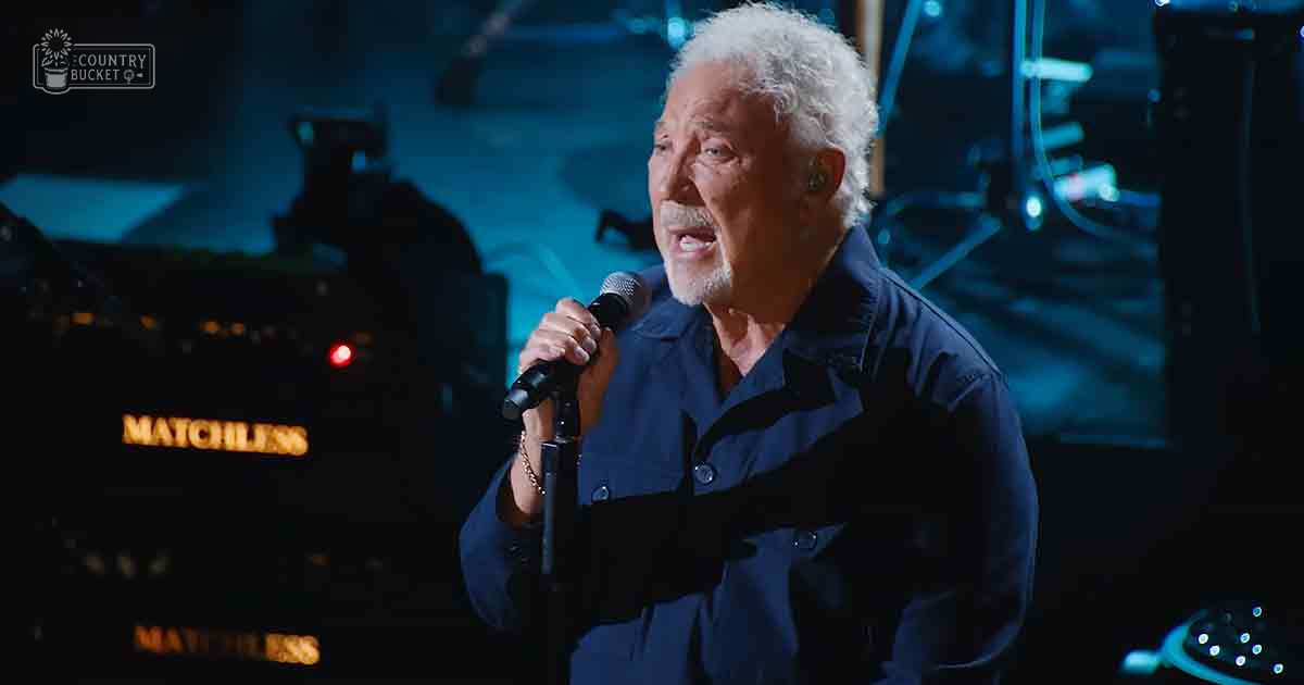 A middle-aged male performer wearing a dark blue button-up shirt with relaxed fit and long sleeves, performing on stage at an indoor concert venue.