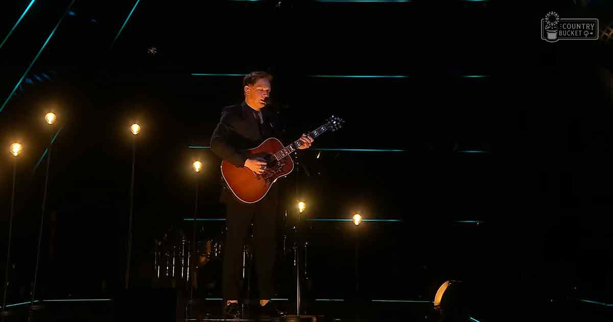 An adult male musician plays a dark-colored, wool-like suit jacket-clad guitar on stage under dim lighting at an evening concert.