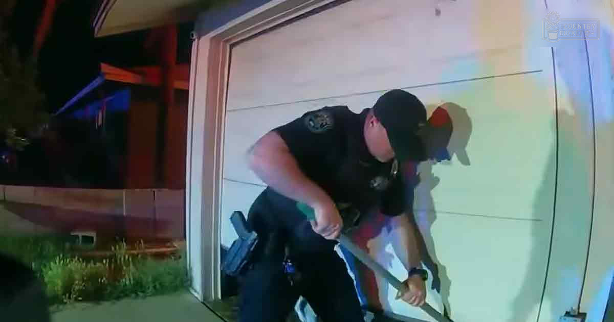 A police officer in a dark uniform stands outside a residential building at night, with a garage door and firearm visible.