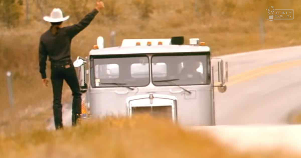 An adult male wearing a black long-sleeve shirt with high collar, dark jeans, cowboy boots, and white cowboy hat stands next to a semi-truck in an open field during early morning or late afternoon light.