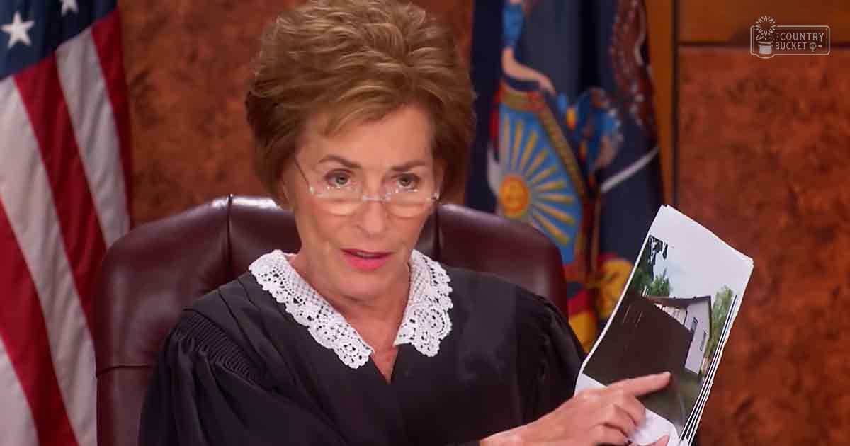 A middle-aged adult female judge in a courtroom setting wearing a black judge's robe with intricate lace detailing, long sleeves with textured cuffs, and glasses. The background includes American flags and another flag with a sun design.