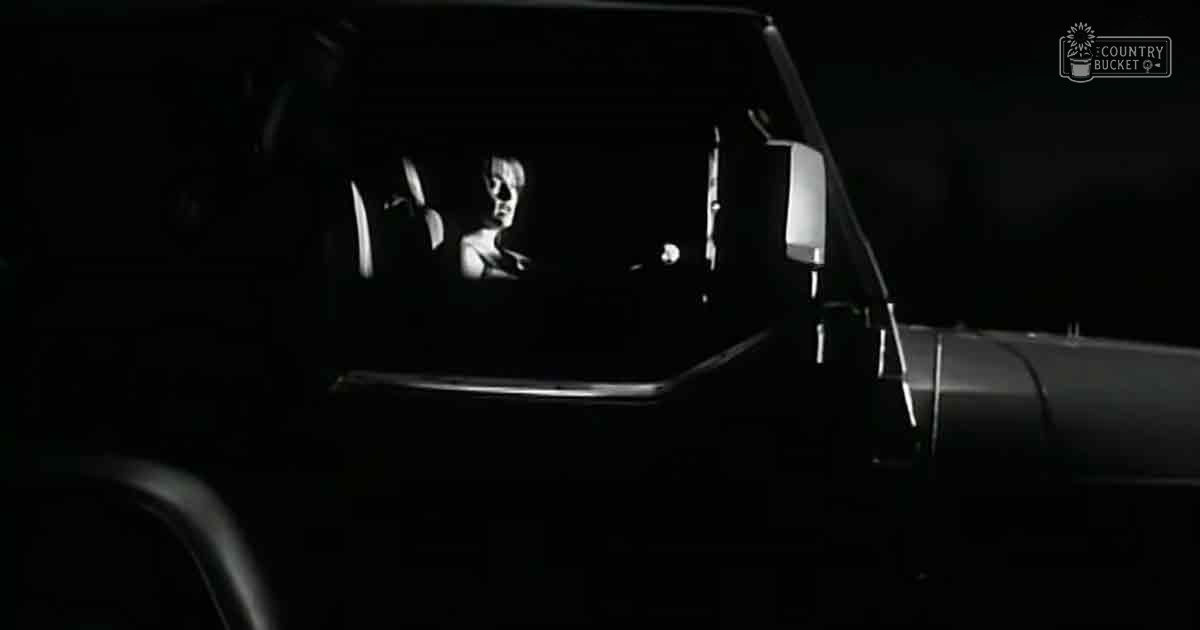 An adult female wearing a dark sleeveless top or dress sits in a partially lit vehicle at night with an urban background