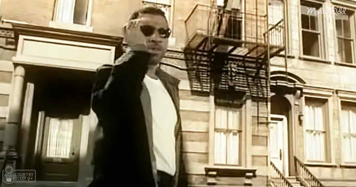 Young adult male wearing black jacket with high collar and dark sunglasses in an urban residential area, possibly New York City.