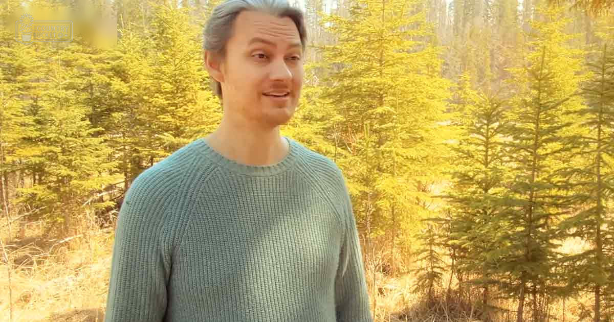 A young adult male wearing a relaxed knit sweater stands in an outdoor forested area with tall trees and sunlight filtering through the foliage, suggesting early morning or late afternoon. The scene is set during autumn.
