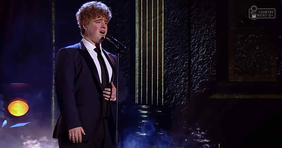 A young adult male in a formal suit performs on stage, wearing a tailored black silk or synthetic suit with a white shirt and black tie, under blue and orange stage lighting.
