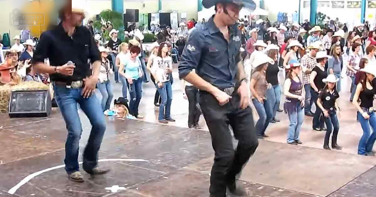 Two men dancing at an outdoor festival with hay bales and a stage setup in the background; various attendees wearing cowboy hats, suggesting a Western theme.