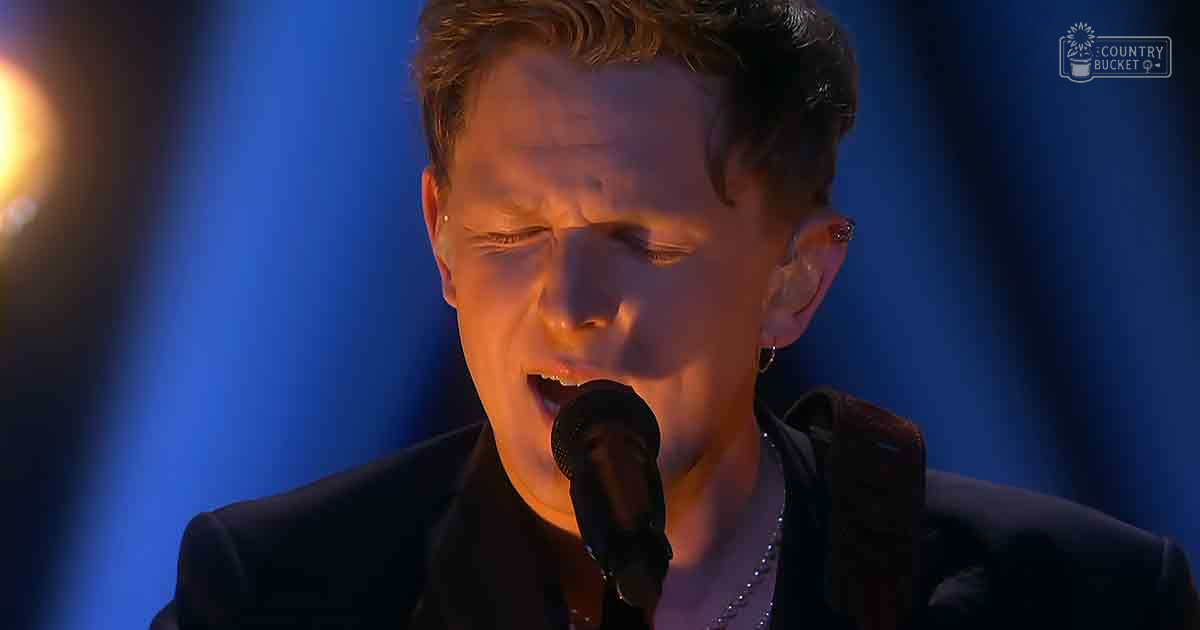 A young adult male performer wearing a dark suit jacket stands on an indoor stage under blue lighting, with a curtain backdrop.