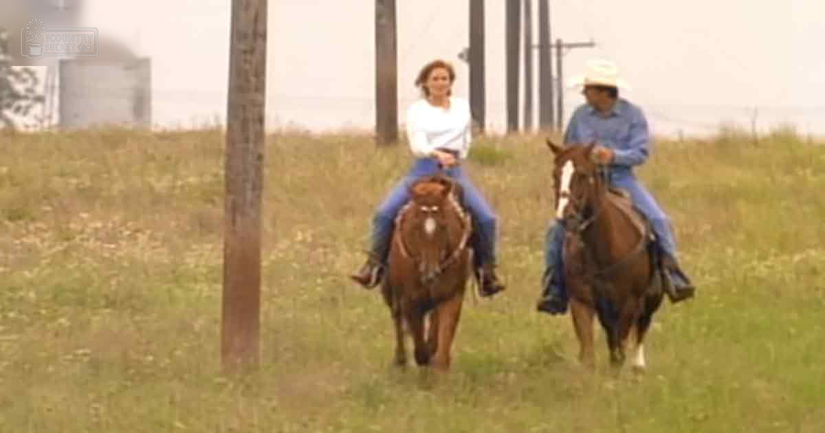 Two individuals riding horses in a rural setting with grassy fields and an industrial silo-like structure in the background under overcast skies.