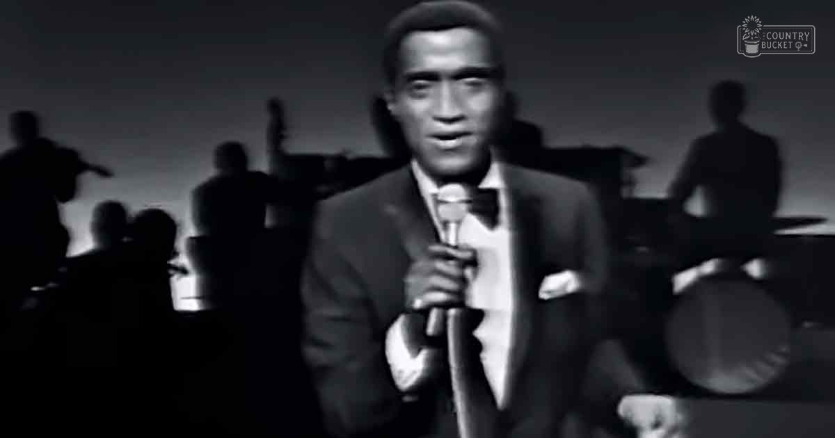 Adult male singer in a dark suit jacket with a white shirt and bow tie performing on stage under dim lighting.