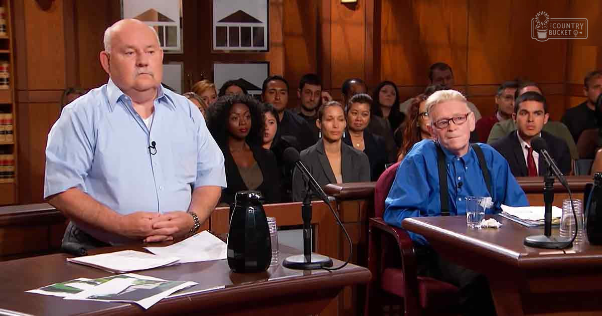 Adult male speaker at podium in courtroom with seated audience and wooden paneling, suggesting a formal indoor setting.