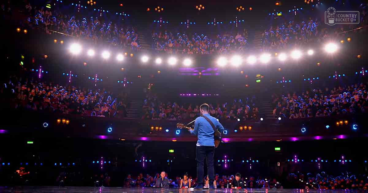 Young adult male musician wearing a blue shirt and brown guitar strap performing on stage at an indoor concert venue with an audience in casual attire.