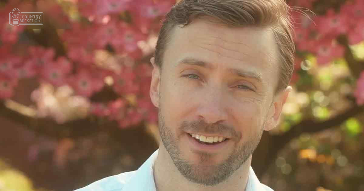 Adult male in a garden with pink blossoms and soft sunlight, wearing a modern casual light blue collared shirt.