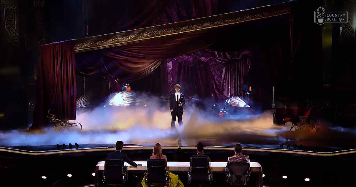 Four seated judges at a long table under dramatic stage lighting in an indoor theater setting with red curtains and gold detailing.