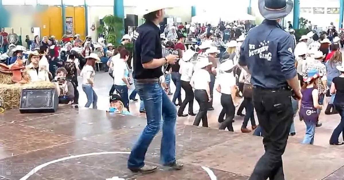 Indoor dance floor scene at a fair or festival with people wearing casual attire, hay bales on one side suggesting Western theme, and various stalls or booths in the background.