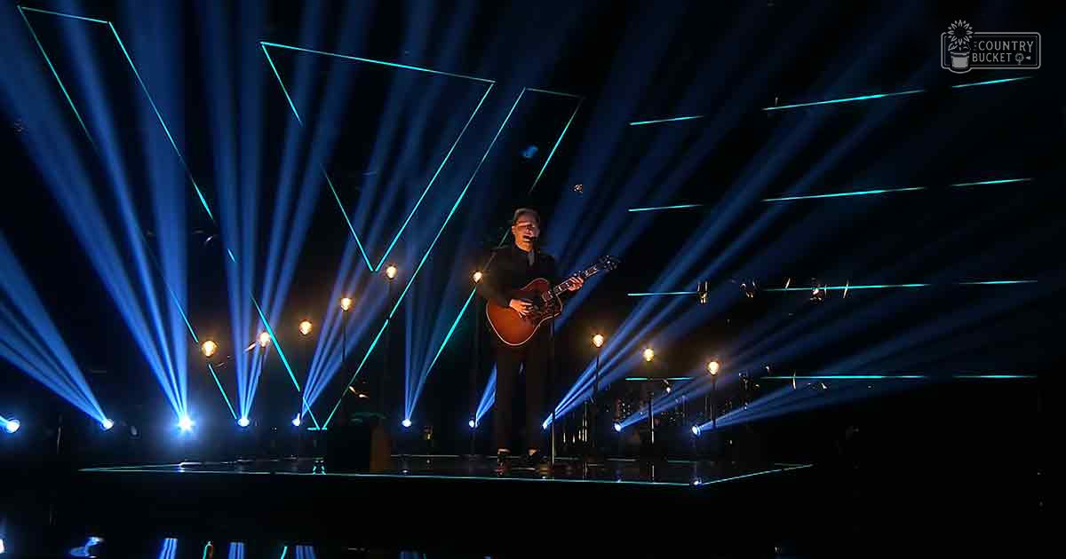 An adult male singer in a dark-colored suit jacket and light shirt performs on stage with dynamic blue and teal lighting, wearing no visible footwear.