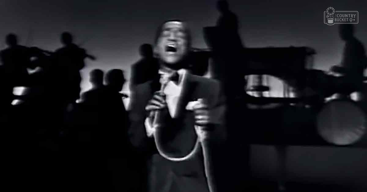 A male adult singer wearing a well-fitted dark suit with a white shirt and light-colored tie, performing in what appears to be a music recording studio setting.