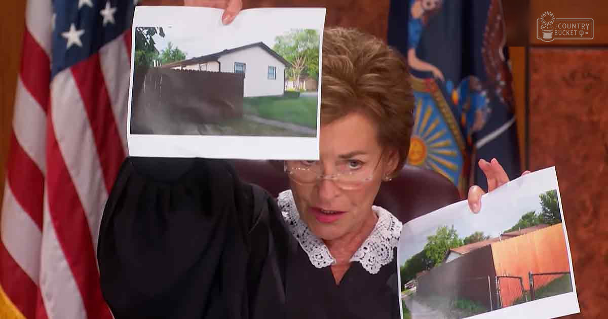 A middle-aged adult female judge wearing a loose, black, solid-color robe with lace-trimmed collar and long sleeves, holding two photographs of houses in an American courtroom setting.